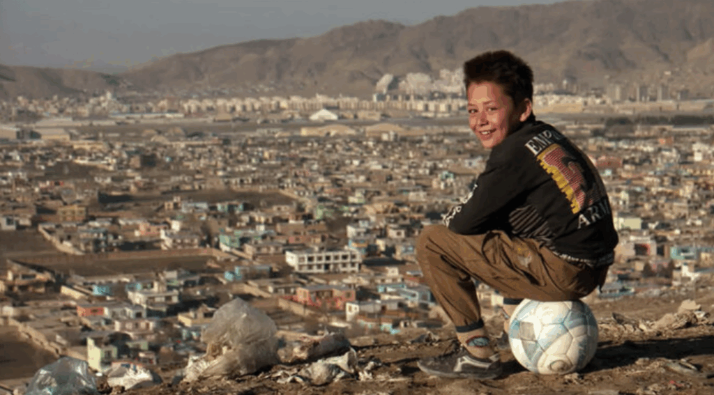 young boy playing soccer. in the background a city in Afghanistan
