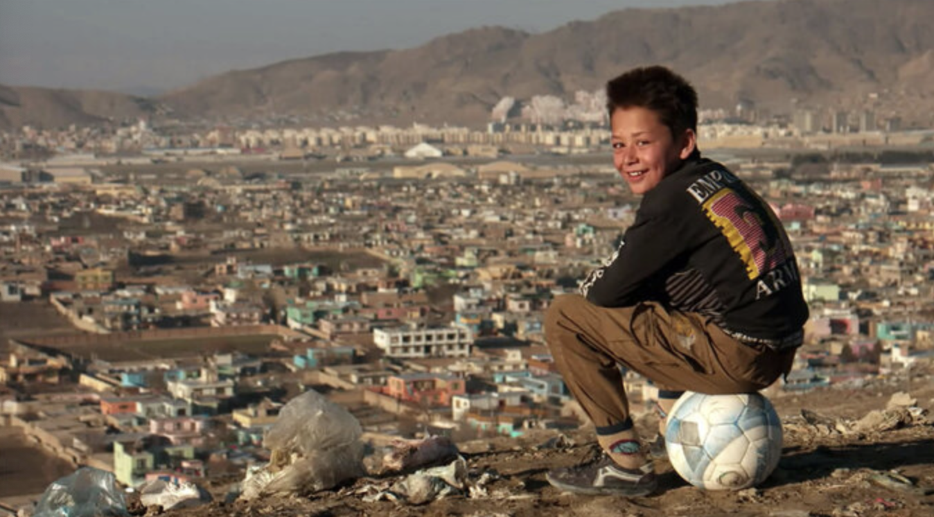 young boy playing soccer. in the background a city in Afghanistan